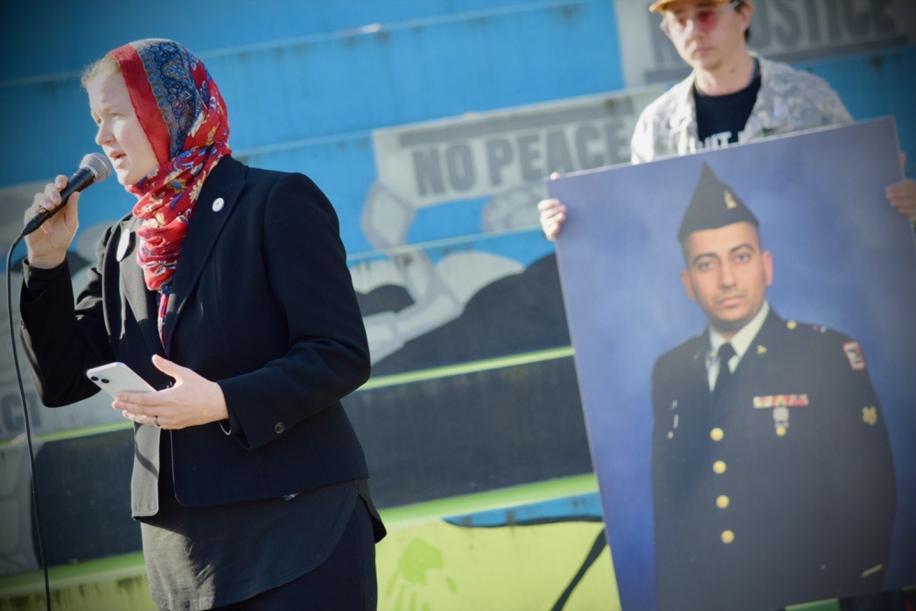 Melissa Chaudhry speaking at an Armistice Day rally, holding a portrait of her husband Zahid in his Army uniform.